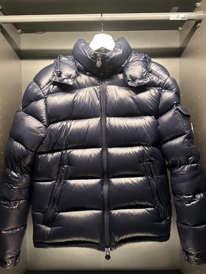 Mörkblå pufferjacka från Moncler - Snygg mörkblå pufferjacka från Moncler med hög krage och avtagbar huva. Jackan har två dragkedjeförsedda fickor framtill och quiltad design med breda kanaler. Tillverkad i glansigt material som ger en exklusiv look. Perfekt för kalla vinterdagar.