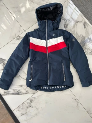 Blå skidjacka från Five Seasons - Snygg marinblå skidjacka med huva från Five Seasons. Jackan har vita och röda detaljer över bröstet, två dragkedjeförsedda fickor och logga på bröstet samt ärmen. Materialet är troligtvis syntet, perfekt för blåsiga dagar. Varm o skön onåka skidor i.Dragkedja framtill och justerbar nederkant.