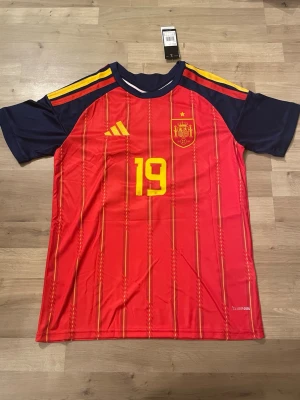 Spanien fotbollströja Lamine Yamal - Röd Spanien fotbollströja från Adidas med gula detaljer, nummer 19 och Lamine Yamal på ryggen. Storlek S 