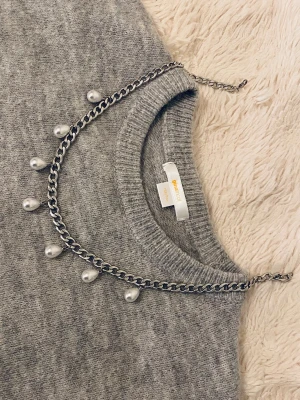 Silverfärgat halsband med pärldroppar - Snyggt halsband i silverfärgad metall med kedja och hängande vita pärldroppar. Perfekt för att ge en cool touch till din outfit. Halsbandet har ett justerbart lås och pärlorna ger en trendig och elegant känsla.