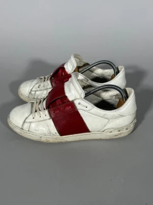 Valentino open - Säljer ett par vita sneakers från Bally med breda röda läderdetaljer över sidan och plösen. Skorna har klassisk låg profil, vita snören och en vit sula. Tillverkade i läder för en clean och stilren look.