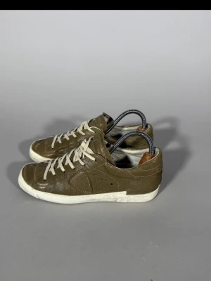 Olivgröna låga sneakers i läder - Snygga olivgröna sneakers i läder med vita sulor och vita snören. Skorna har en klassisk låg profil och diskreta sömmar. Insidan har en brun detalj vid hälen. Perfekta för dig som gillar stilrena och tidlösa sneakers.