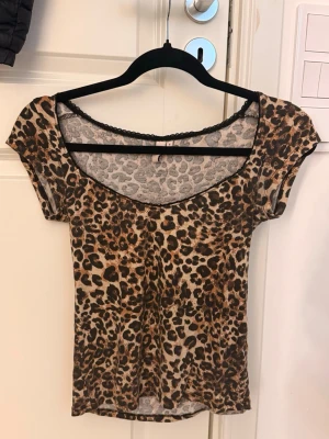 Leopardmönstrad topp med spetskant - Snygg topp med leopardmönster och rundad urringning. Toppen har korta ärmar och en tunn spetskant längs halsringningen för en extra detalj. Materialet känns mjukt och stretchigt, perfekt för att skapa en cool look.