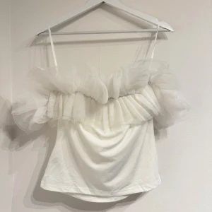 Vit offshoulder topp med volang - Supersöt vit topp med tunna axelband och stora, fluffiga volanger i tyll längs offshoulder-kragen. Perfekt för dig som vill sticka ut med en romantisk och trendig look. Toppen har en mjuk, draperad passform och är riktigt snygg till både jeans och kjol.