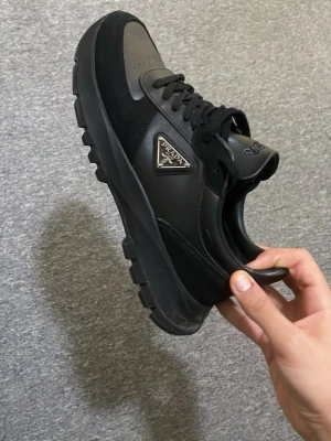 Svarta Prada sneakers i läder och nylon - Snygga svarta sneakers från Prada med chunky sula och ikonisk metallogo på sidan. Skorna är gjorda i en mix av läder och nylon, har svarta skosnören och en modern, sportig look. Perfekta för dig som gillar exklusiva och trendiga detaljer.