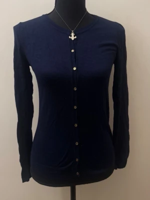 Mörkblå klassisk kofta med knappar - En stilren mörkblå kofta med rund halsringning och långa ärmar. Koftan har silverfärgade knappar framtill och är tillverkad i ett tunt, mjukt material som sitter figurnära. Perfekt att matcha med jeans eller kjol.