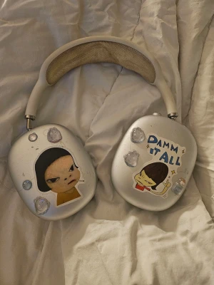 Airpodmax - Snygga vita over-ear hörlurar med vadderad bygel och unika stickers på kåporna, bland annat en tecknad figur och texten 'DAMN IT ALL'. Hörlurarna har en modern, rundad design och ser riktigt bekväma ut. Perfekt för dig som vill sticka ut med din stil.