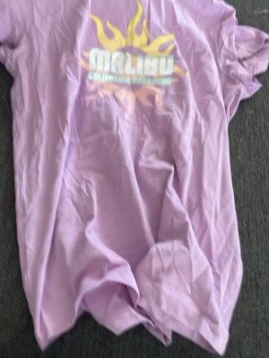 Lila Malibu t-shirt från Reaction - Ljuslila t-shirt från Reaction med tryck av en gul sol och texten 'Malibu California dreaming' på bröstet. Klassisk rund hals och korta ärmar. Perfekt för dig som gillar färgglada och somriga plagg.