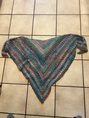 Flerfärgad virkad trekantssjal - Säljer en handvirkad trekantssjal i mjukt garn med färger i blått, grönt, rosa och grått. Sjalen har ett luftigt mönster och passar perfekt att svepa runt axlarna för en cool och avslappnad look. Perfekt accessoar för att lyfta din outfit.
