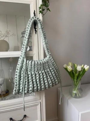 Ljusgrön virkad handväska - Trendig ljusgrön handväska med bred axelrem och chunky virkad design. Väskan är gjord i ett mjukt, tjockt garn som ger en unik och modern look. Perfekt accessoar för dig som vill sticka ut med en cool och personlig stil.