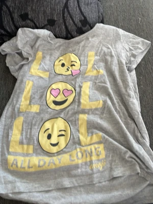 Grå emoji t-shirt med tryck - Grå t-shirt från emoji med stort tryck av gula emojis och texten 'LOL ALL DAY LONG' i gult framtill. T-shirten har korta ärmar och är gjord i mjuk bomull, perfekt för en chill och lekfull stil.