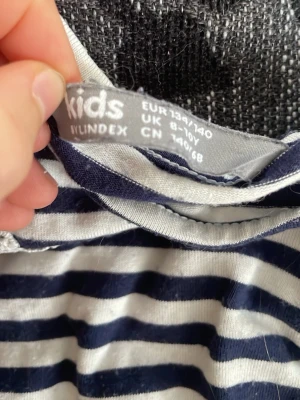Randig långärmad topp från Lindex Kids - Snygg långärmad topp från Lindex Kids i marinblått och vitt med horisontella ränder. Toppen har spetsdetaljer på axlarna och är tillverkad i mjuk bomull. Perfekt för en avslappnad och trendig look.