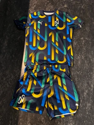 Färgglatt Neymar x Puma matchställ - Säljer ett Neymar x Puma matchställ med t-shirt och shorts. Setet har ett färgstarkt mönster i blått, grönt och gult med stora siffror 10 över hela plagget. Tillverkat i lätt och snabbtorkande material, perfekt för fotboll eller träning.
