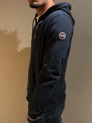 Mörkblå huvtröja från Colmar - Snygg mörkblå hoodie från Colmar med dragkedja framtill och huva med dragsko. Mjukt material och ribbade muddar vid ärmslut och nederkant. Colmar-logga på vänster ärm ger en sportig vibe. Perfekt till jeans eller joggers.