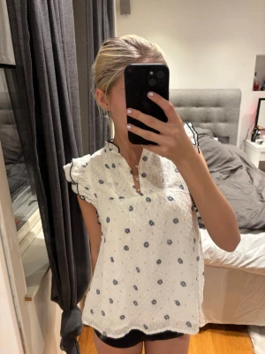 Vit blommig blus med volangdetaljer - Supersöt vit blus med små blå blommor och volangdetaljer längs ärmhålen. Blusen har v-ringning med knapp och svarta broderade kanter på volangerna. Luftig och lätt i materialet, perfekt för sommaren.