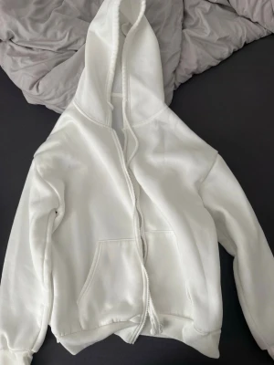 Vit hoodie med dragkedja från SHEIN - Säljer en vit hoodie från SHEIN i storlek S. Hoodien har dragkedja framtill, stor huva och två fickor på magen. Tillverkad i 100% polyester vilket gör den mjuk och skön. Perfekt för chill dagar eller när du vill ha en enkel och clean look.