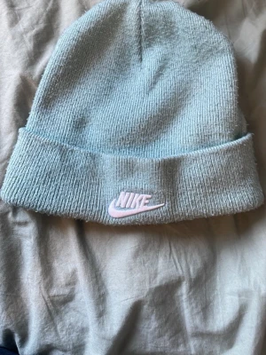Ljusblå ribbad mössa från Nike - Säljer en ljusblå ribbstickad mössa från Nike med uppvikt kant och broderad logga framtill i vitt och rosa. Perfekt för dig som vill ha en enkel men snygg accessoar till hösten eller vintern. Materialet känns mjukt och bekvämt.