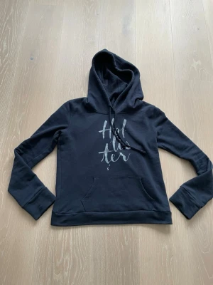 Svart hoodie från Hollister - Snygg svart hoodie från Hollister med tryckt logga framtill i vitt. Klassisk modell med huva och dragsnören samt en stor magficka. Tillverkad i mjukt bomullsmaterial och har långa ärmar. Perfekt för en avslappnad och trendig look.