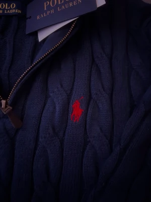 Mörkblå kabelstickad tröja Polo Ralph Lauren - Snygg mörkblå kabelstickad tröja från Polo Ralph Lauren med halv dragkedja och röd broderad logga på bröstet. Tröjan har ribbade muddar och hög krage, perfekt för lager på lager. Klassisk och tidlös design i mjukt material.