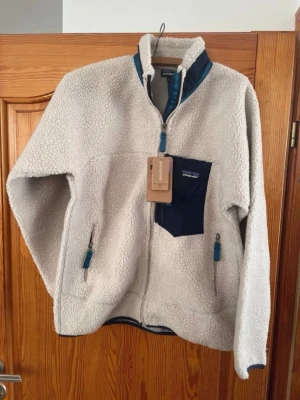 Beige teddyjacka från Patagonia - Mysig beige teddyjacka från Patagonia med blå detaljer. Jackan har hög krage, dragkedja framtill, tre fickor med dragkedja och en kontrasterande bröstficka. Perfekt för kalla dagar och har en avslappnad passform.