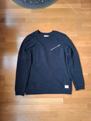 Svart sweatshirt Jack & Jones Core - Svart sweatshirt från Jack & Jones Core med rund halsringning, snygg bröstficka med dragkedja. Tröjan har ribbade muddar och armbågar.. Perfekt för en avslappnad och stilren look. Storlek L med är passar till M-S.