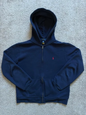 Mörkblå hoodie från Polo Ralph Lauren - Mörkblå hoodie med dragkedja från Polo Ralph Lauren. Klassisk design med huva, två fickor framtill och den ikoniska röda logotypen broderad på bröstet. Tillverkad i mjukt bomullsmaterial som är skönt att bära. Perfekt för en avslappnad och stilren look.