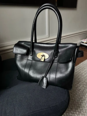 Mulberry väska i läder med gulddetalj - Stilren svart handväska i äkta läder med två rejäla handtag och snygg guldfärgad vridknäppe framtill. Klassisk rektangulär form och diskreta sömmar ger en elegant look. Perfekt för dig som gillar tidlös design och vill ha plats för det viktigaste.