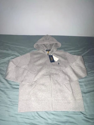 Grå zip hoodie från Polo Ralph Lauren - Snygg grå hoodie med dragkedja från Polo Ralph Lauren. Klassisk design med huva, två fickor framtill och den ikoniska blå loggan broderad på bröstet. Tillverkad i mjuk bomullsblandning som känns skön mot huden. Perfekt för en avslappnad och stilren look.