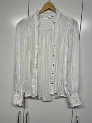 Vit blus med volangkrage från Co couture  - Stilren vit blus från Co couture med diskreta ränder, volangkrage och knappar framtill. Blusen har långa ärmar med breda smockade muddar och lätt puff vid axlarna. Tillverkad i ett tunt, mjukt tyg som ger en luftig känsla.