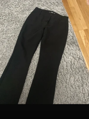 Svarta kostymbyxor med raka ben - Snygga svarta kostymbyxor med klassisk rak passform och hällor för skärp. Byxorna har diskreta fickor och är perfekta för en clean och stilren look. Materialet känns mjukt och följsamt, vilket gör dem bekväma att bära hela dagen.