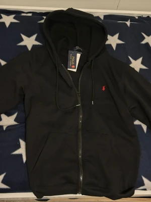 Svart hoodie från Polo Ralph Lauren - Svart hoodie med dragkedja från Polo Ralph Lauren. Klassisk design med huva och dragsnören, två fickor framtill och den ikoniska röda logotypen broderad på bröstet. Perfekt för en avslappnad och stilren look.