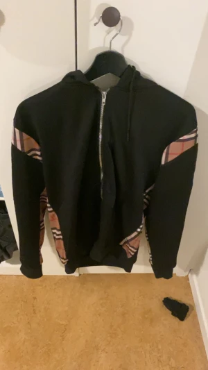 Svart hoodie burberry hoodie - Säljer en svart hoodie med dragkedja och huva. Ärmarna har snygga rutiga paneler i beige, rött och svart som ger en unik look. Hoodien har snörning i huvan och är tillverkad i mjukt sweatshirtmaterial. Perfekt för dig som vill ha en streetwear vibe. Pris går att diskutera 