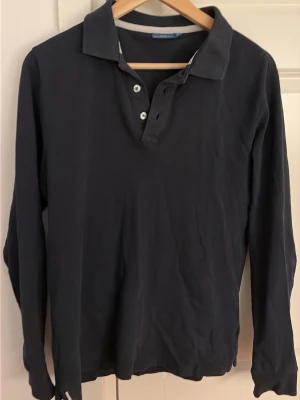 Mörkblå långärmad piké från C&S - Snygg mörkblå långärmad pikétröja från Cotton & Silk med klassisk krage och tre knappar framtill. Tillverkad i mjukt bomullsmaterial och har en enkel, stilren design som passar till det mesta.