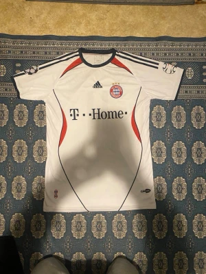 Bayern München matchtröja Podolski 11 L - Säljer en vit Bayern München matchtröja från Adidas med Podolski och nummer 11 på ryggen. Tröjan har svarta och röda detaljer, klubbmärke på bröstet och T-Home som sponsor. Perfekt för dig som älskar fotboll och vill ha en klassisk supportertröja.