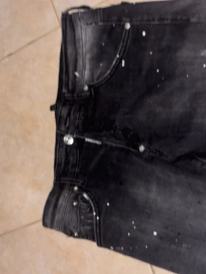 Svarta jeans med stänkdetaljer - Säljer ett par svarta jeans med coola vita stänkdetaljer och klassisk femficksmodell. Jeansen har en något tvättad look och silverfärgade knappar. Perfekta för dig som vill ha en edgy och trendig stil.