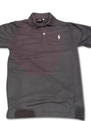 Mörkgrå pikétröja från Polo Ralph Lauren - Storlek S men passar även M. Klassisk mörkgrå pikétröja från Polo Ralph Lauren med vit broderad logga på bröstet. Tröjan har korta ärmar, krage och knappar framtill. Perfekt till en avslappnad men ändå stilren look.