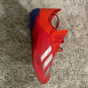 Röda Adidas fotbollsskor med dobbar - Snygga röda fotbollsskor från Adidas med klassiska vita ränder på sidan. Skorna har en strömlinjeformad design, snörning och metall-dobbar för bra grepp på planen. Ovandelen är i syntetmaterial och sulan har blå och genomskinliga detaljer.