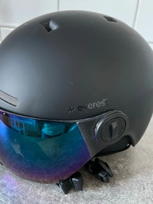 Svart skid/snowboardhjälm Everest - Säljer en matt svart skid- eller snowboardhjälm från Everest med integrerat visir i blått och lila spegelglas. Hjälmen har ventilationshål och justerbart spänne under hakan. Perfekt för dig som vill ha både skydd och stil i backen.