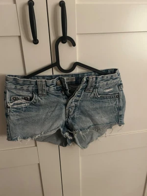 Blå jeansshorts med rå kant - Säljer ett par ljusblå jeansshorts med råa, fransiga kanter och slitna detaljer. Klassisk femficksmodell med bälteshällor och knappgylf. Perfekta för varma dagar och en avslappnad look.