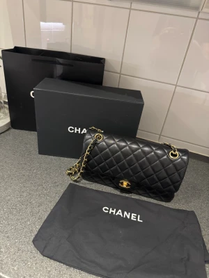 Chanel svart handväska - Säljer en ikonisk svart quiltad handväska med klassisk kedjerem i guld och vridlås med logga. Väskan är tillverkad i läder och har en tidlös, elegant design som passar perfekt till alla outfits. Dustbag och originalbox medföljer.