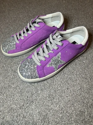 Lila glitter sneakers från Golden Goose - Snygga lila sneakers från Golden Goose med silverglittrig tå och stjärna på sidan. Skorna har gråa skosnören och vit sula. Materialet är en mix av mocka och glitterdetaljer som ger en unik look. Vänligen kontakta mig innan köp!