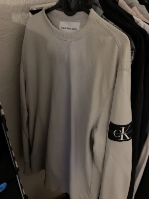 vit sweatshirt från Calvin Klein - vit sweatshirt från Calvin Klein Jeans med rund halsringning och ribbade muddar. På vänster ärm sitter en svart patch med Calvin Klein-loggan. Tröjan har en clean och stilren look, perfekt för en avslappnad stil.