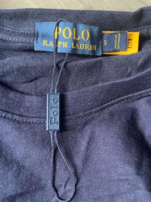 Mörkblå t-shirt från Polo Ralph Lauren - Snygg mörkblå t-shirt från Polo Ralph Lauren med klassisk gul logga broderad på bröstet. T-shirten har rund hals och korta ärmar, tillverkad i mjuk bomull för en skön känsla. Perfekt till jeans eller shorts för en clean look.