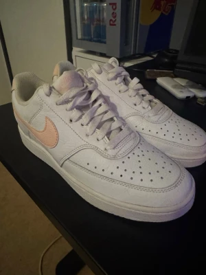 Vita Nike sneakers med rosa detaljer - Snygga vita Nike sneakers med ljusrosa swoosh och detaljer på häl och plös. Skorna har klassisk låg profil, perforerad tåbox för ventilation och vita skosnören. Tillverkade i läder med textilfoder. Perfekta för en clean och fräsch look.
