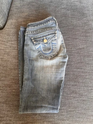 Blå bootcut jeans med kontrastsömmar - Säljer ett par blå bootcut jeans med slitningar och kontrastsömmar. Jeansen har två bakfickor med lock och dekorativa sömmar samt knappdetaljer. Klassisk femficksmodell i denim med snygg tvätt.