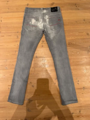 Jacob Choën Jeans - Snygga grå jeans i slim fit-modell med röda sömmar på bakfickorna. Jeansen har klassisk femficksdesign och är tillverkade i ett mjukt denimtyg. Perfekta för dig som gillar en stilren och modern look.