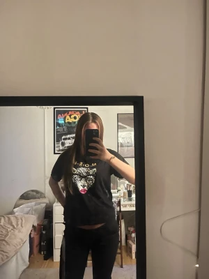 Svart t-shirt med leopardtryck - Cool svart t-shirt med tryck av en leopard med glasögon och rosa läppar samt texten M.O.R.E. Trycket är i vitt, guld och rosa. T-shirten har korta ärmar och en klassisk passform. Perfekt för dig som vill sticka ut lite extra.