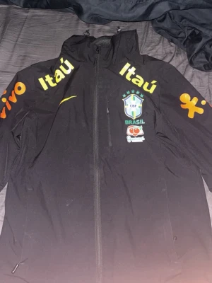 Svart Brasilien Nike träningsjacka - Säljer en svart träningsjacka från Nike med Brasilien CBF-logga på bröstet. Jackan har huva, hel dragkedja och tryck i gult och grönt med sponsorer som Itaú och Vivo. Perfekt för fotbollsträning eller som sportig streetwear.