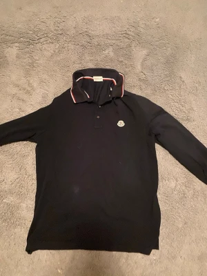 Svart långärmad piké från Moncler - Snygg svart långärmad pikétröja från Moncler med klassisk krage och vita samt röda detaljer på kragen. Tröjan har Moncler-logga på bröstet och knappar vid halsen. Perfekt för en stilren och avslappnad look.
