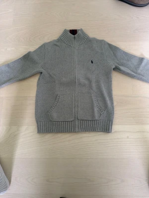 Grå stickad ziptröja med fickor - Snygg grå stickad tröja med dragkedja framtill och två fickor. Tröjan har ribbade muddar vid ärmslut och nederkant samt en liten broderad logga på bröstet. Hög krage med randig detalj på insidan ger extra stilpoäng.